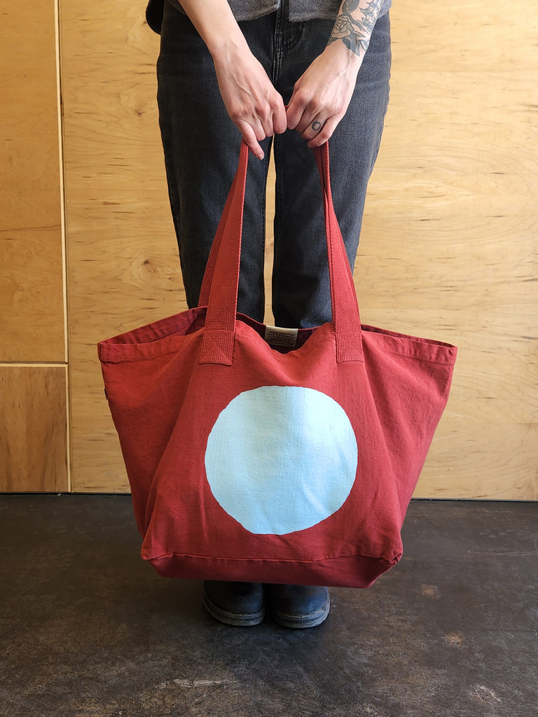 A cranberry red tote bag with a pale blue large circle printed on it.
